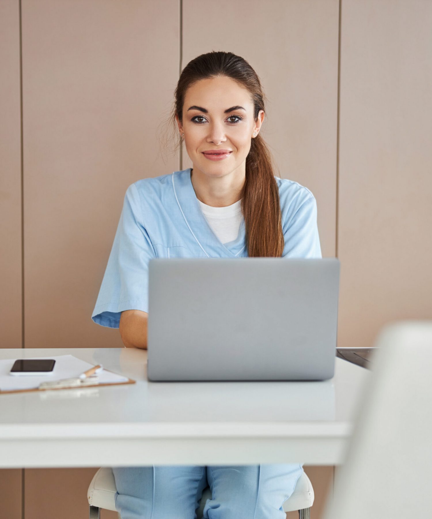 Attractive young lady in doctor scrubs sitting at the table with laptop and looking at the camera